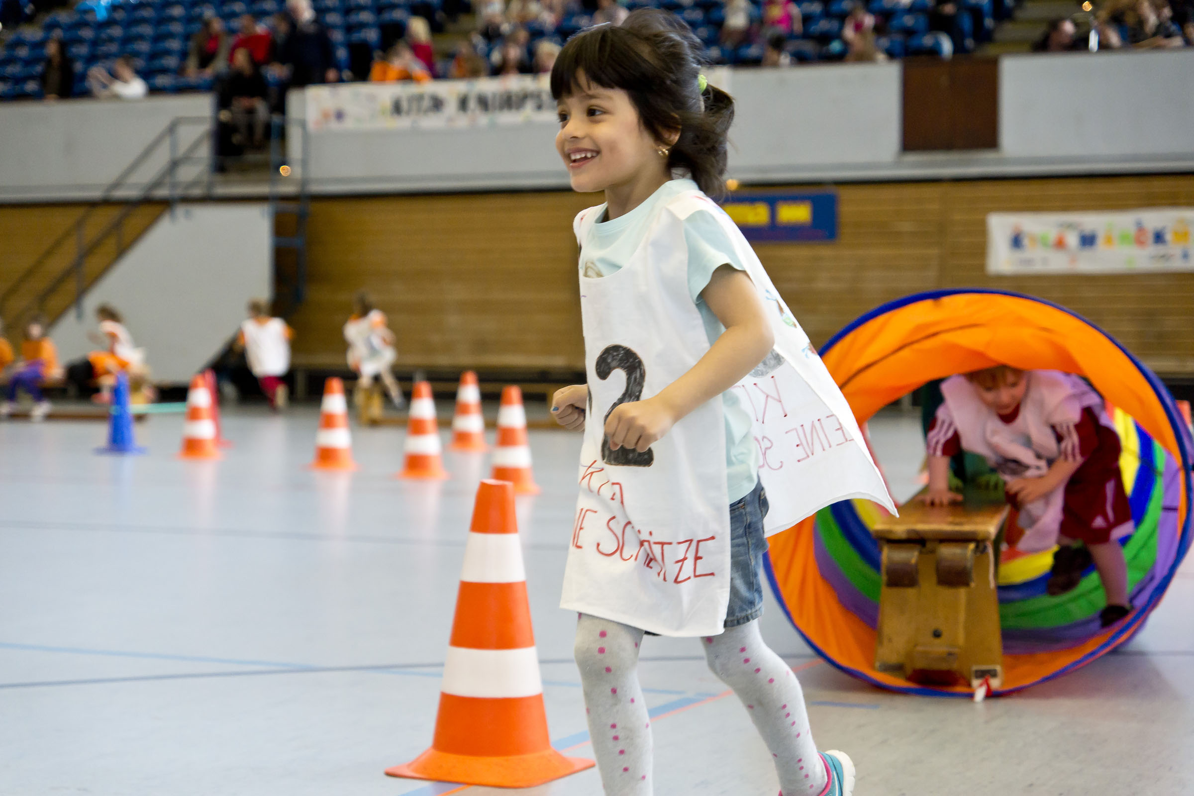 Mehrere Kita-Kinder laufen gemeinsam unter einem gelben Schwungtuch durch eine Sporthalle