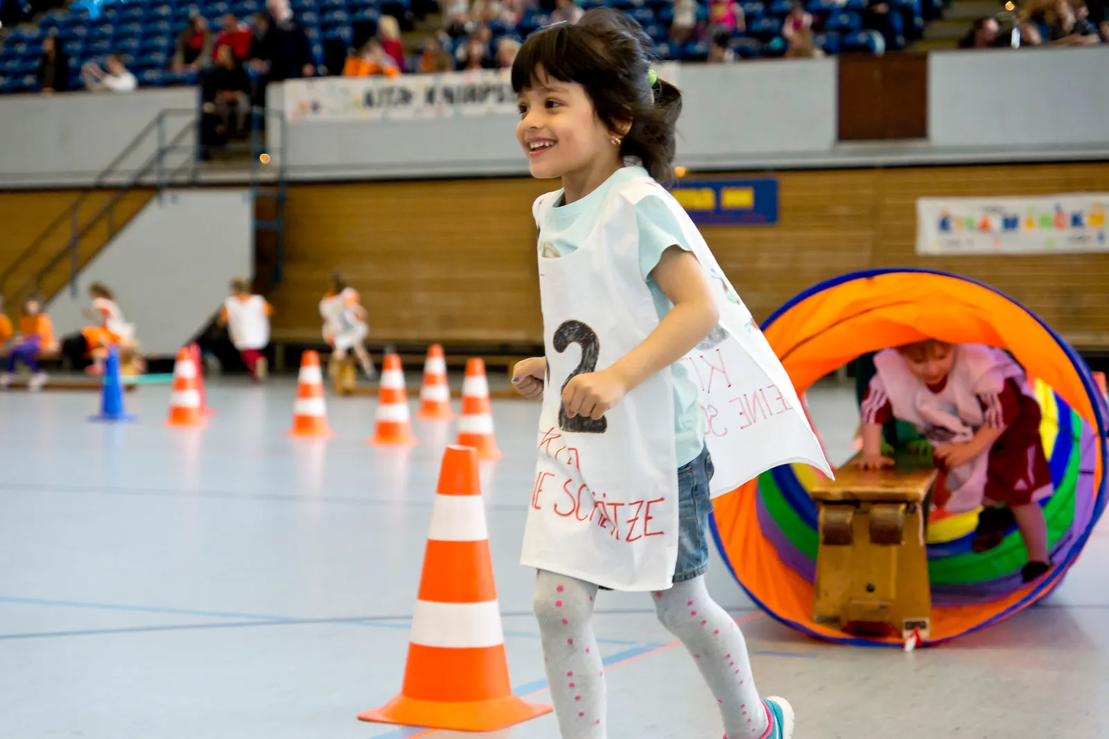 Mehrere Kita-Kinder laufen gemeinsam unter einem gelben Schwungtuch durch eine Sporthalle